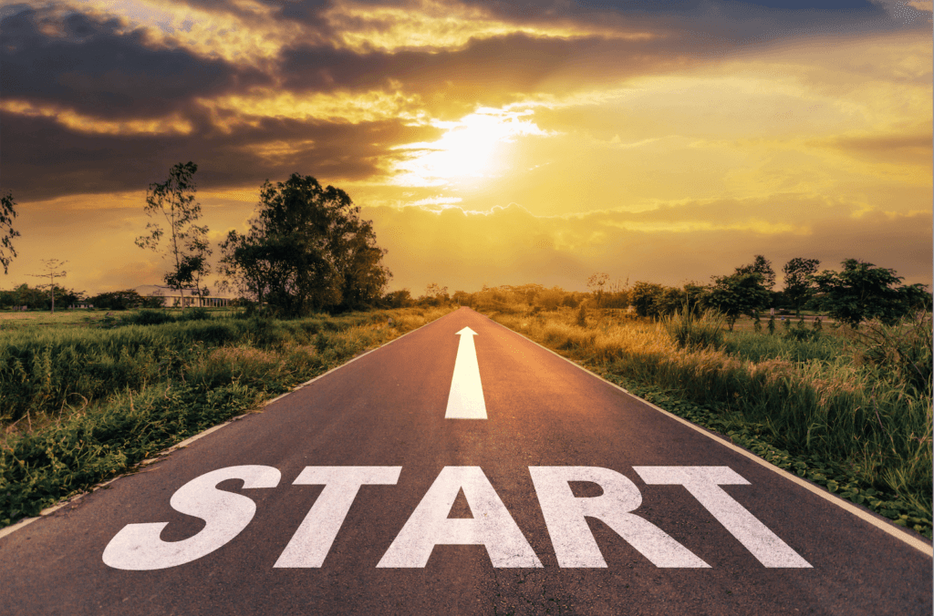 A paved road stretches into the distance under a dramatic sky with a bright sunset, casting a golden glow. The word "START" is prominently painted in large white letters at the beginning of the road. Lush green fields and trees line both sides of the road, creating a serene and inviting atmosphere.